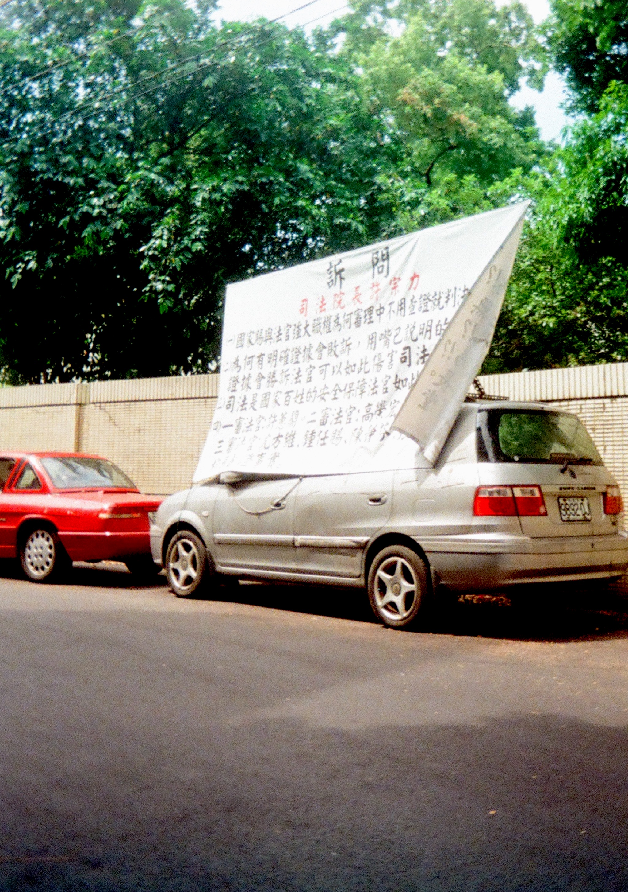 A parked car with a banner hanging on top of it, covering the driver's side. Banner reads some complaints about the judiciary systems.